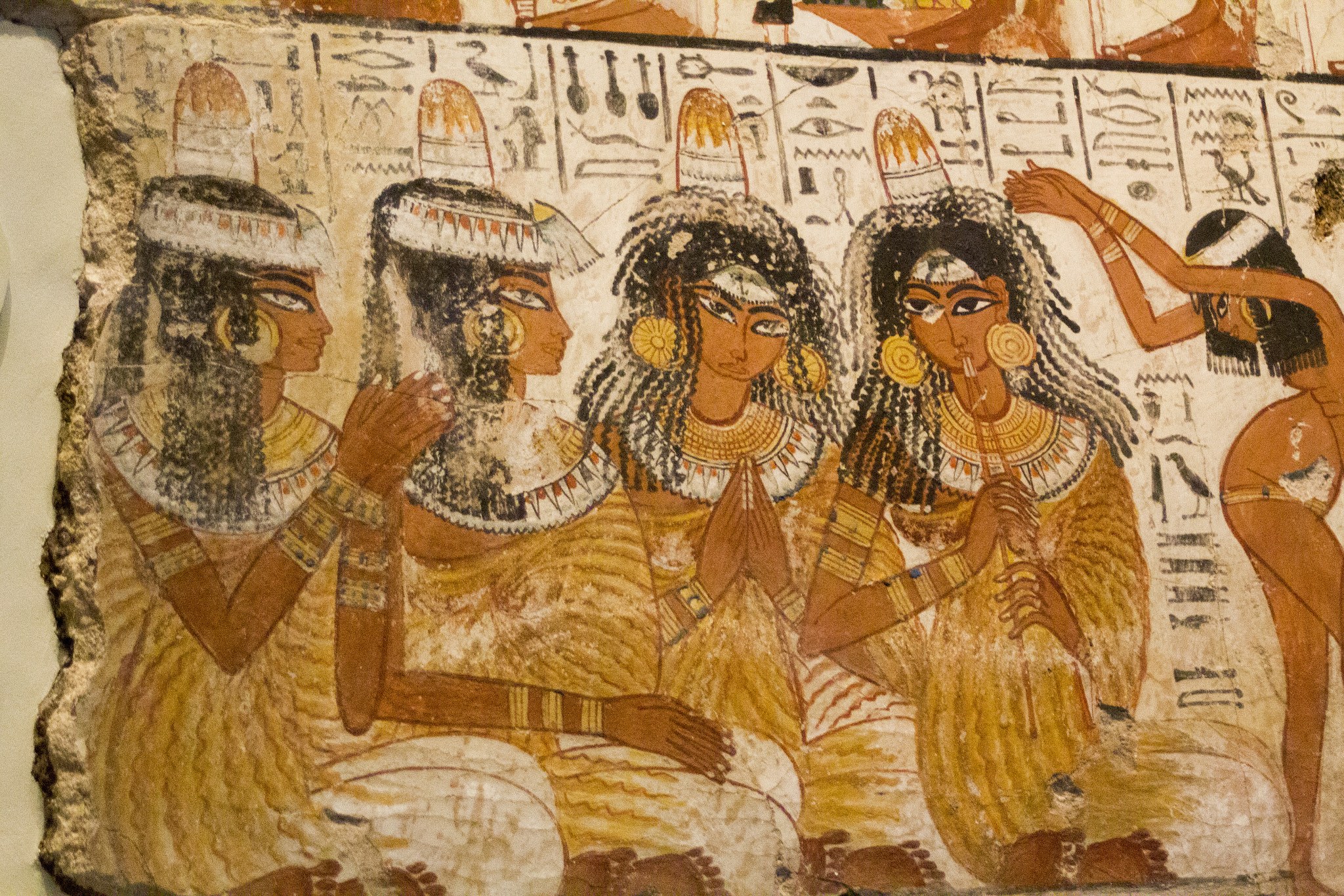 Fragment of ancient Egyptian tomb wall painting showing four seated figures in profile with gold jewelry and hieroglyphic text above.