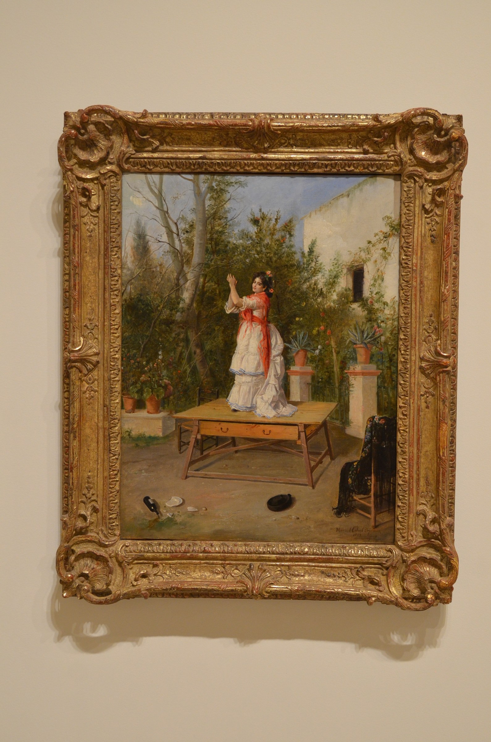 Woman in white petticoat and red sash dances on a wooden table in a sun-lit garden courtyard, arms raised, observed by a seated woman.