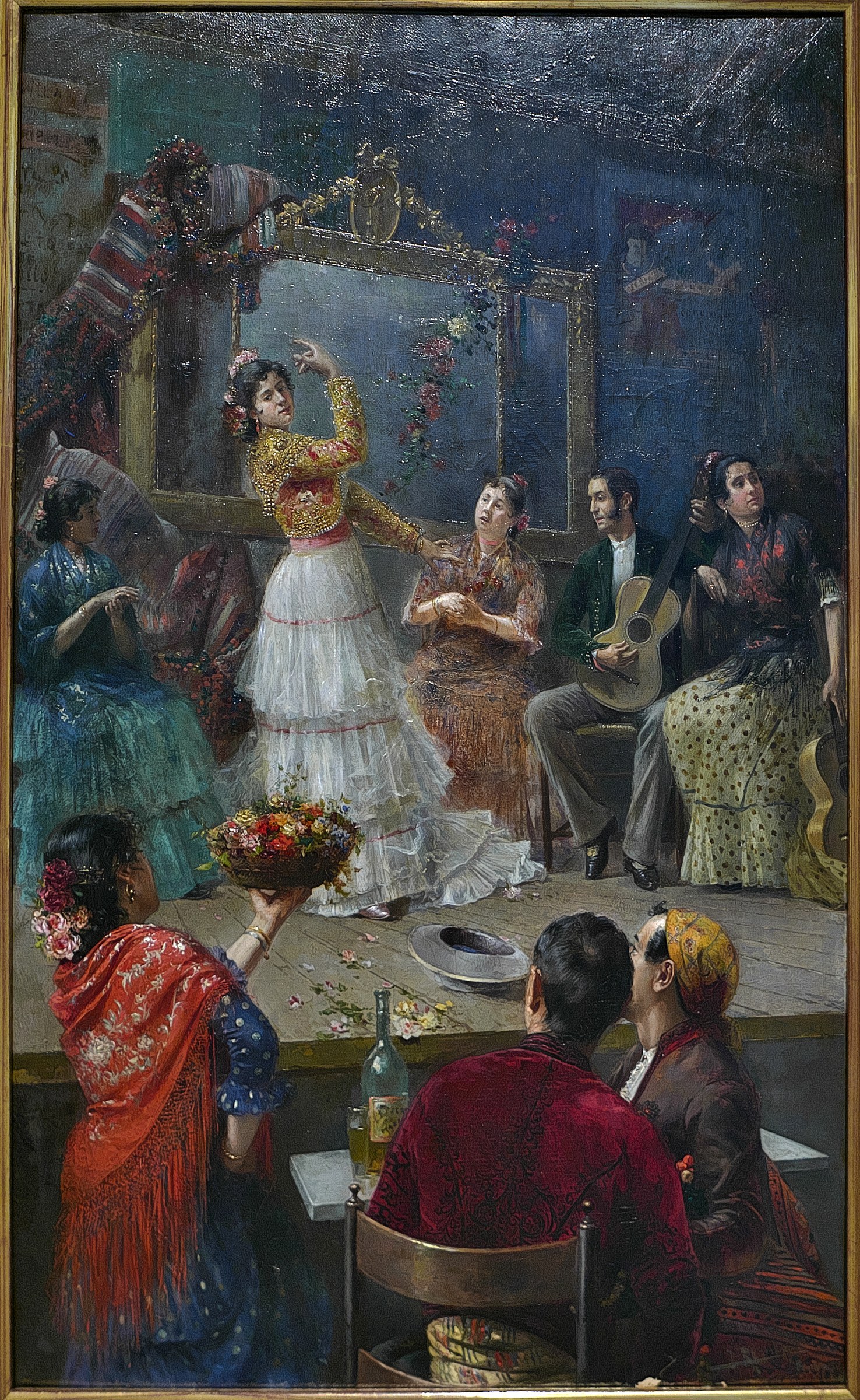 A woman in a yellow jacket and white skirt dances in the center of a candlelit room, one arm raised in a flamenco pose, surrounded by seated spectators and a