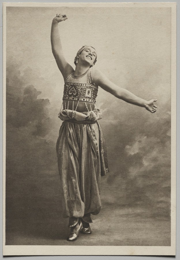 Male dancer in ornate tunic and trousers, one arm raised, body extended in arabesque against clouded background.