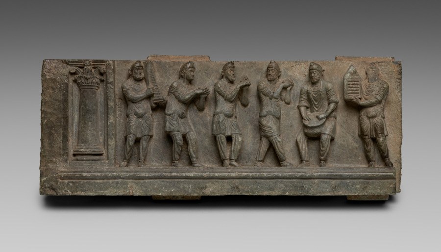 Ancient schist relief showing six Phrygian figures: three dancing with raised arms, two playing musical instruments, one holding an unidentified object.