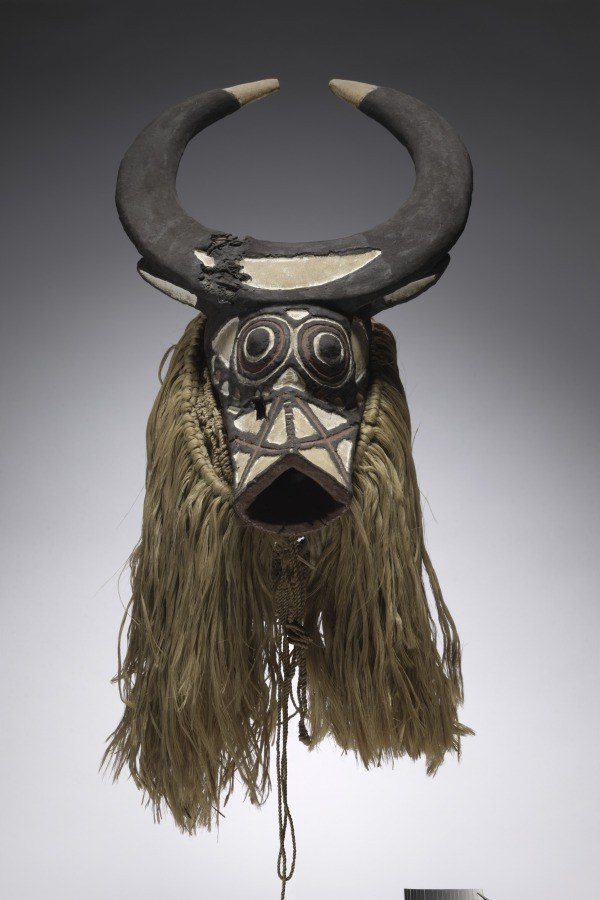 Dark wood mask with upward-curving horns, carved animal muzzle, inlaid eyes, and dense straw-colored fiber beard hanging below.