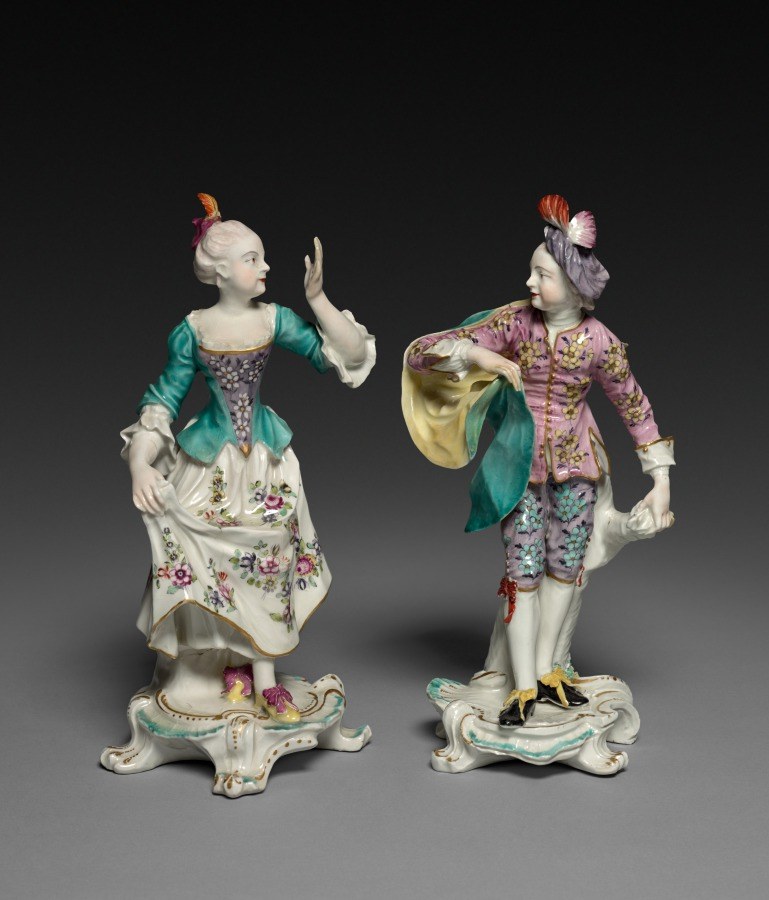 Two porcelain figurines in 18th-century theatrical dress stand on rococo bases, gesturing toward each other in a moment of courtly social interaction.