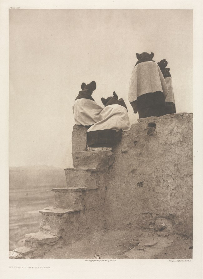 Three wrapped figures stand on ancient stone steps, gazing across a misty landscape; they are spectators watching a ceremony or dance.