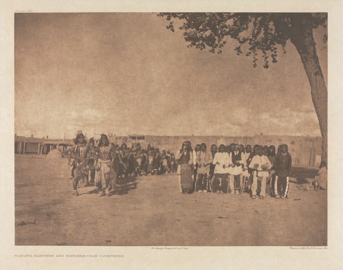 Large gathering of Pueblo people in a plaza under a tree, wearing traditional dress and blankets, for a Tablita ceremony.