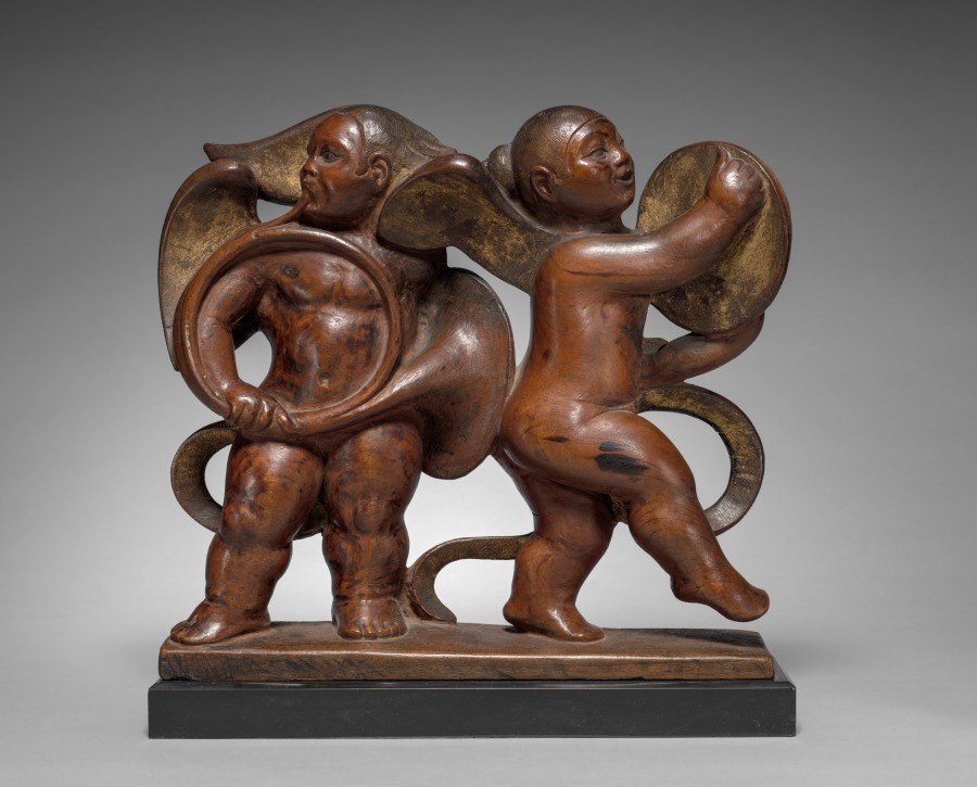 Wooden relief of two muscular putti in dynamic poses with ornamental scrollwork behind them.