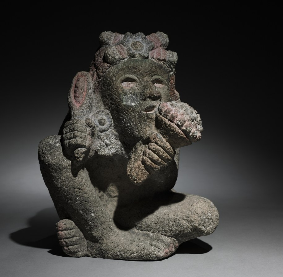 Stone sculpture of a seated Aztec deity with an elaborate flower crown, holding objects, with maize cobs across the shoulders.