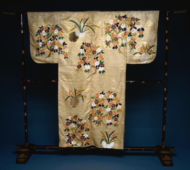 Cream silk nō costume embroidered with purple and pink cherry and iris blossoms and gold leaf, displayed flat on a wooden stand.