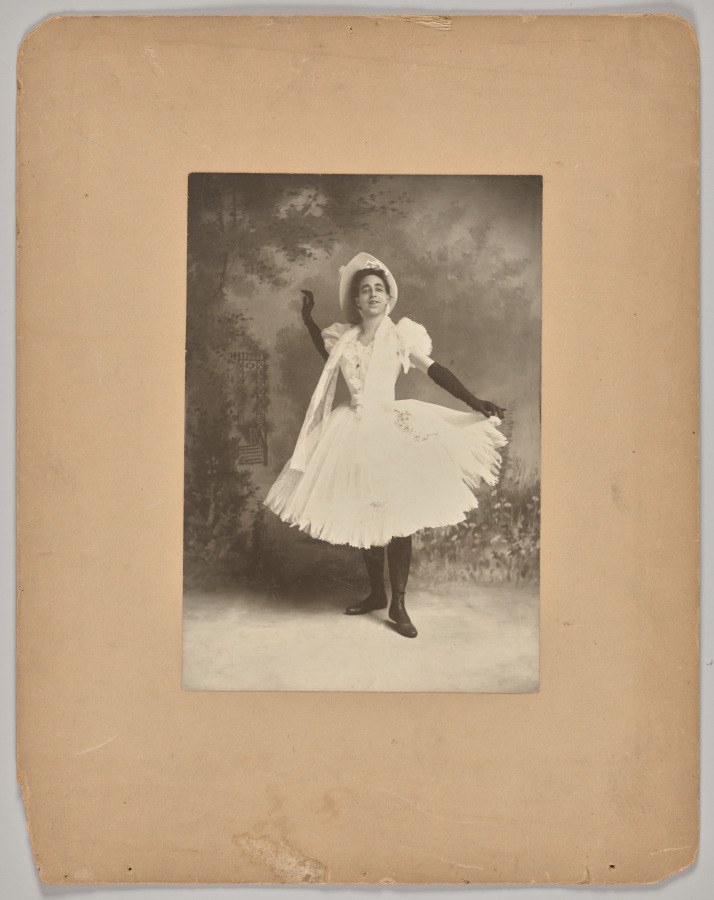 A male dancer in a white Romantic ballet costume—pleated skirt, bonnet, bodice—poses with one arm raised, mid-gesture, against a painted backdrop.