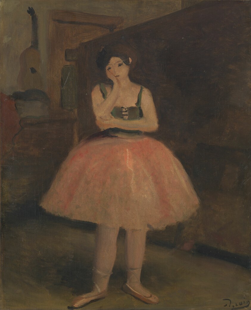 Young female ballet dancer in pink tutu and pointe shoes standing alone in a dim studio, one hand raised to her face, gazing downward pensively.
