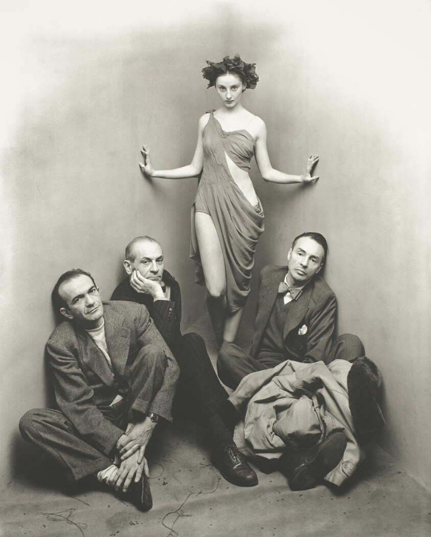 Five figures in a studio: a woman in classical drapery stands elevated with arms open; four men in business suits crouch below, gazing upward.