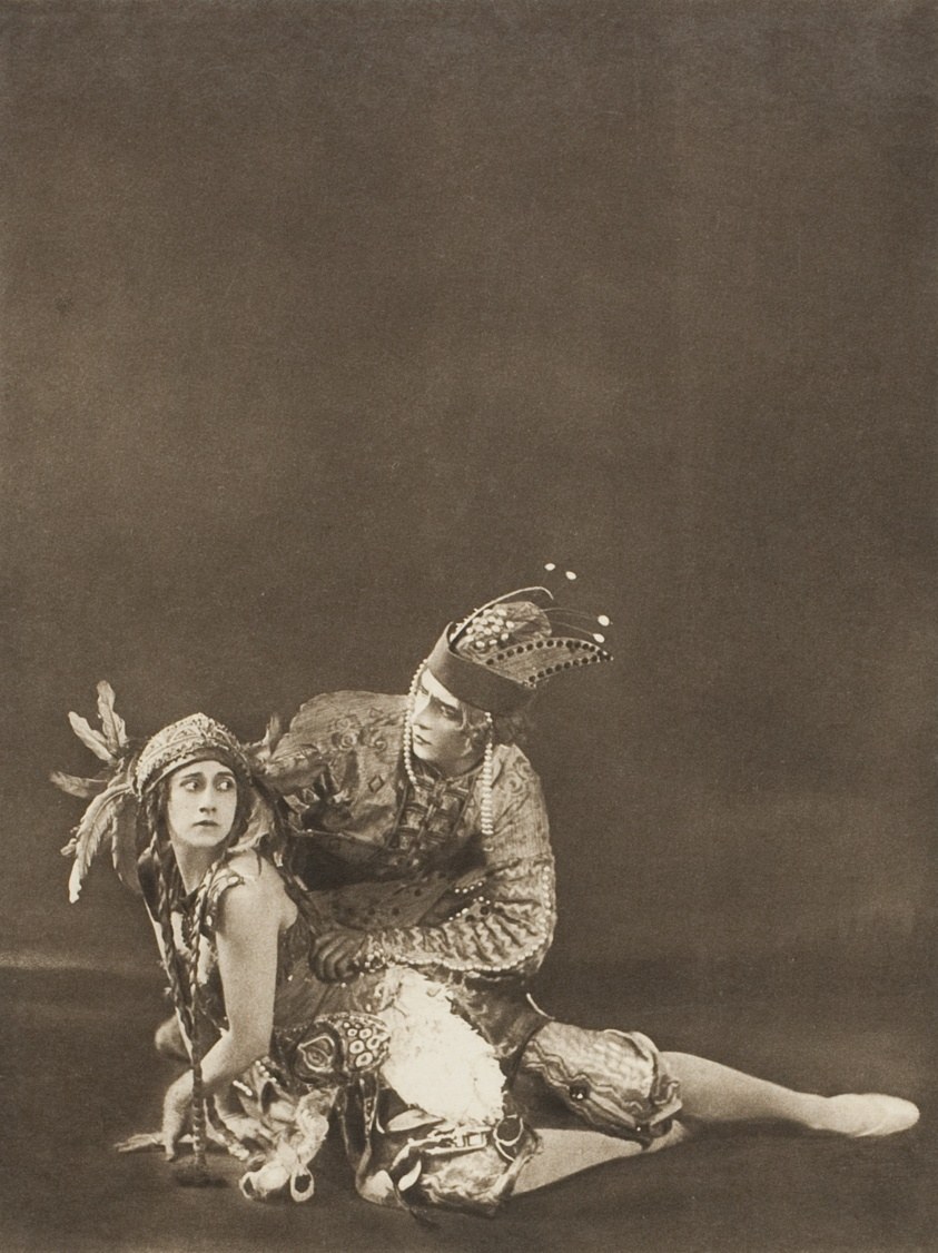 Male dancer kneels beside seated female dancer in elaborate Russian folk-inspired costumes with tall headdresses and jeweled tunics against dark background.