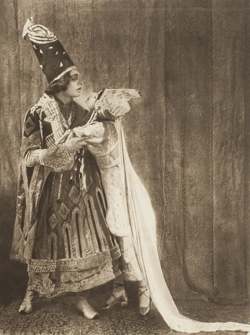 A dancer in ornate dark tunic and white train, crowned with tall plumed headdress, stands in formal profile against draped backdrop.