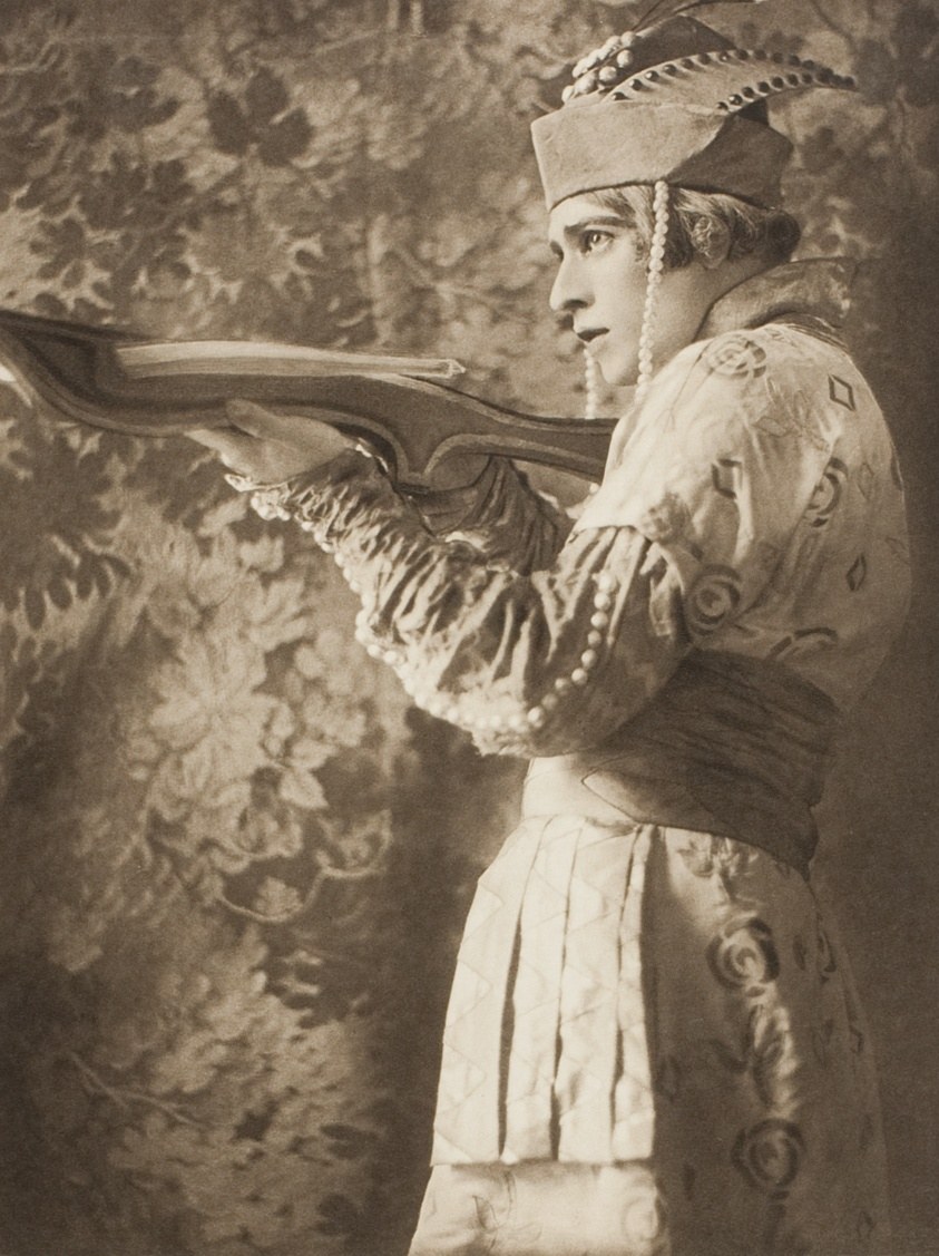 Male dancer in profile in white tunic and feathered headdress, holding a large curved wing prop, against a damask backdrop.