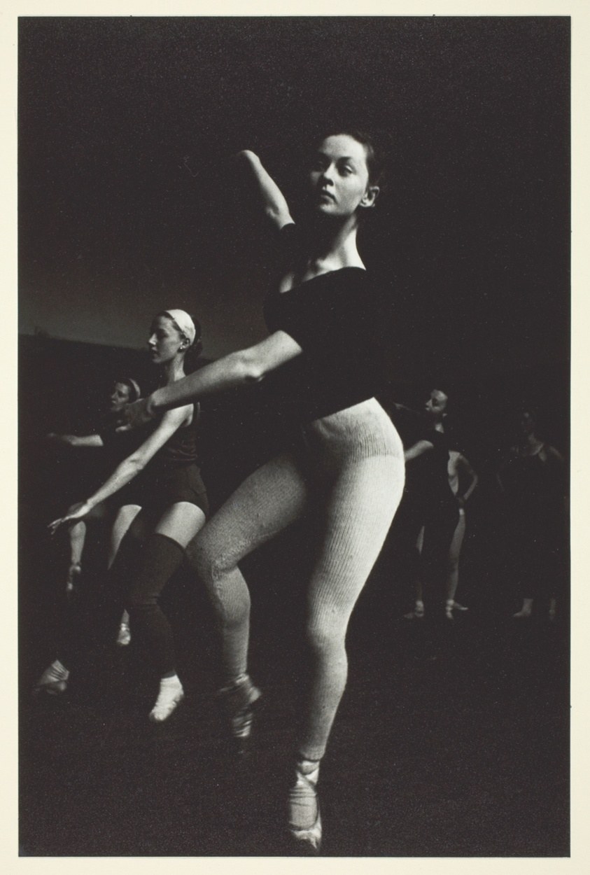 Male ballet dancer in dark leotard holds a vertical relevé with raised arm; three younger dancers watch from the shadowed background.