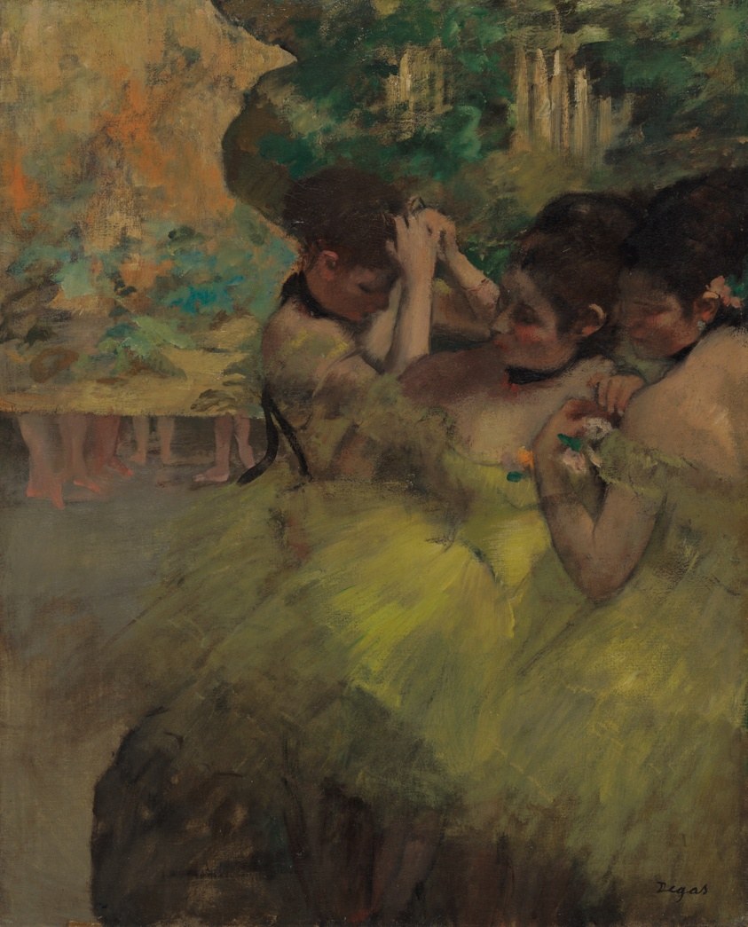 Three young women in pale yellow tutus rest backstage, one adjusting her hair, two seated close together.