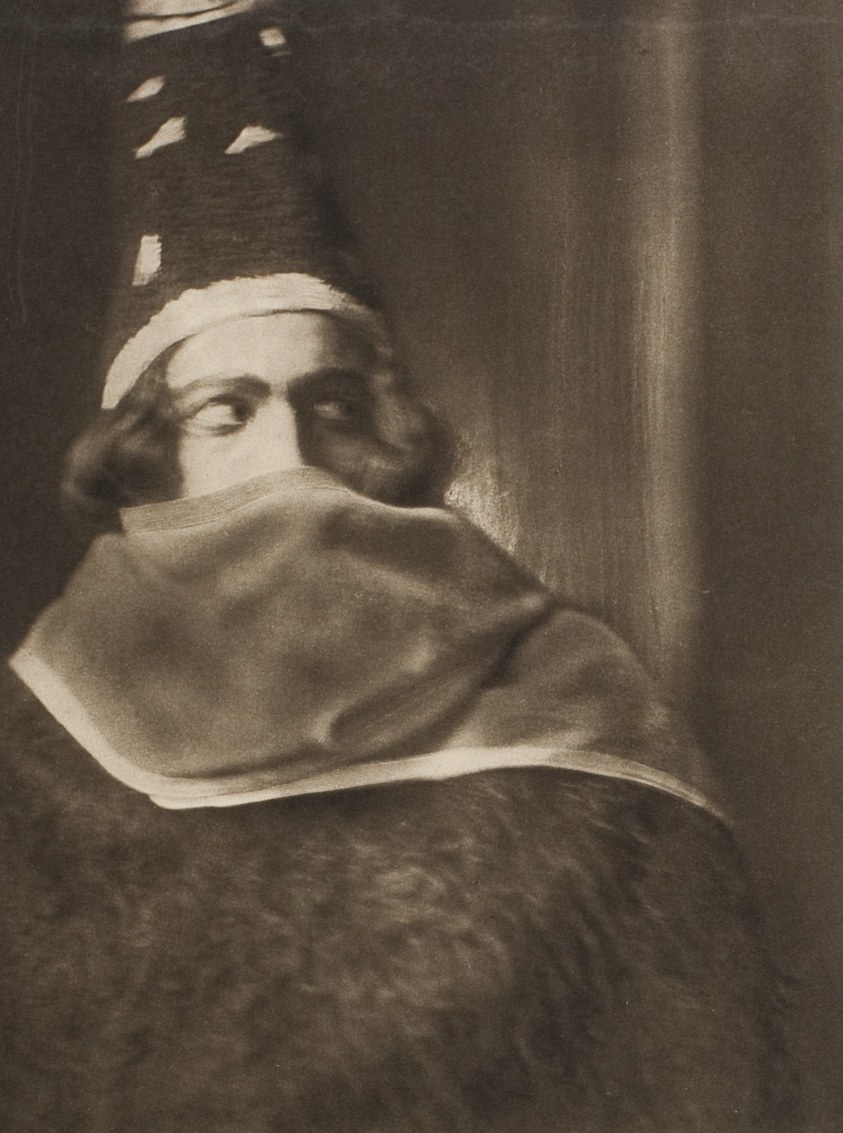 Male dancer in turban and draped robes stands in contemplative stillness, gazing downward, in a sepia-toned photographic portrait.