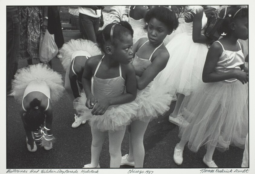 Young Black children in white ballet tutus and pointe shoes stand in a crowded outdoor parade setting, holding classical ballet posture.