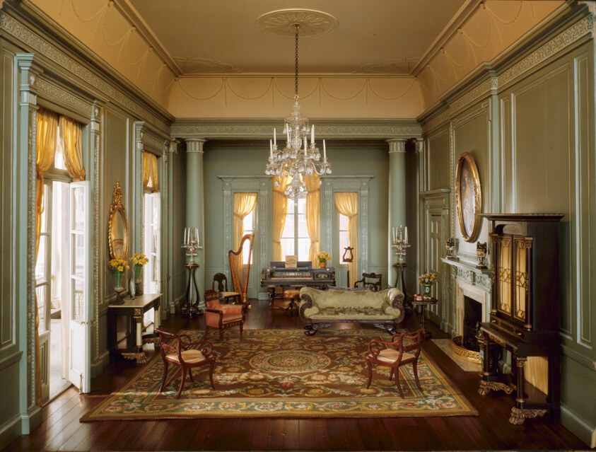 Empty South Carolina ballroom with green walls, tall windows with yellow drapes, chandelier, and mahogany chairs arranged along perimeter.