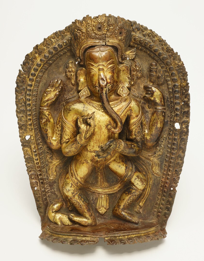 Gilt bronze relief of four-armed Ganesha in frontal pose, wearing crown and jewelry, with a small rat at his feet, against ornate patterned background.