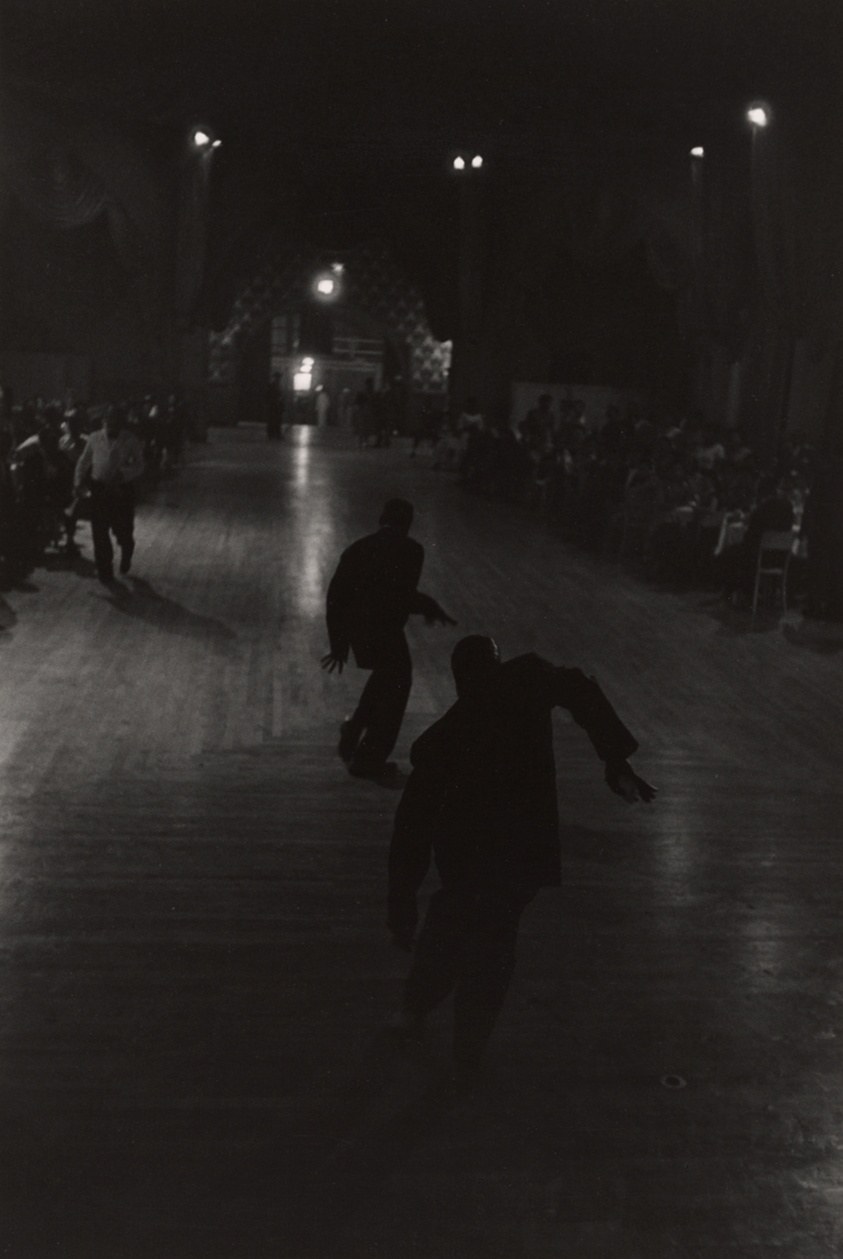 Two men in dark suits stride across a wet New York street at night, illuminated by overhead lamps, with onlookers visible in shadow.