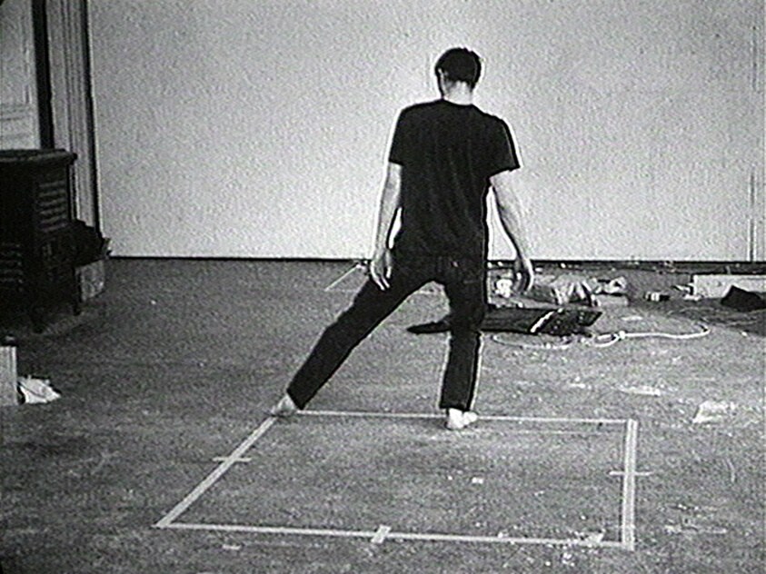 Black-and-white video of a solitary figure in dark clothing straddling a white square marked on the floor, arms at sides, in a wide-legged stance.