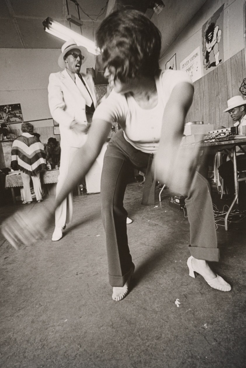 Shirtless male dancer twists and reaches mid-movement in an industrial studio; female dancer behind; observers in white suits stand aside.