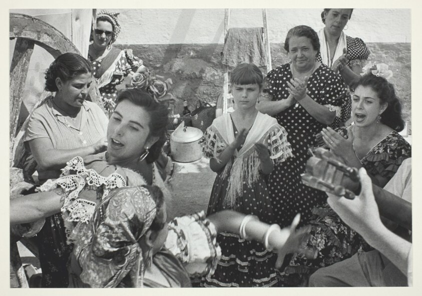 Women and a boy gathered outdoors in a circle, animated and dancing, with stone walls behind them.