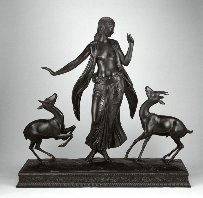 Bronze sculpture of a classical female figure in draped garment, flanked by two graceful gazelles in profile stance on a rectangular plinth.