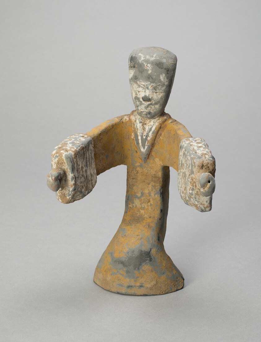 Gray ceramic figurine of a standing woman in a long robe with raised arms, head simplified, on a conical base.