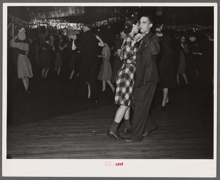 Black-and-white photograph of a crowded dance hall with couples dancing in close holds on a wooden floor, spectators visible in the background.