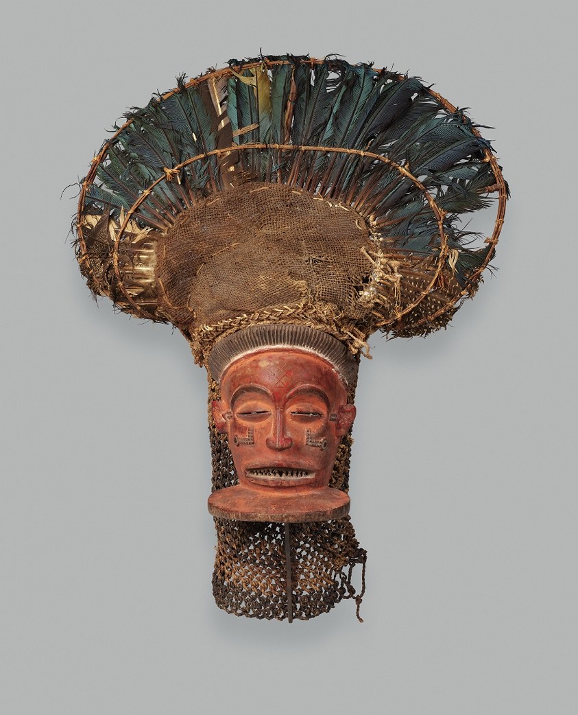Wooden mask with terracotta-red face, metal neck rings, and towering headdress of woven raffia trimmed with dark teal and white feathers.