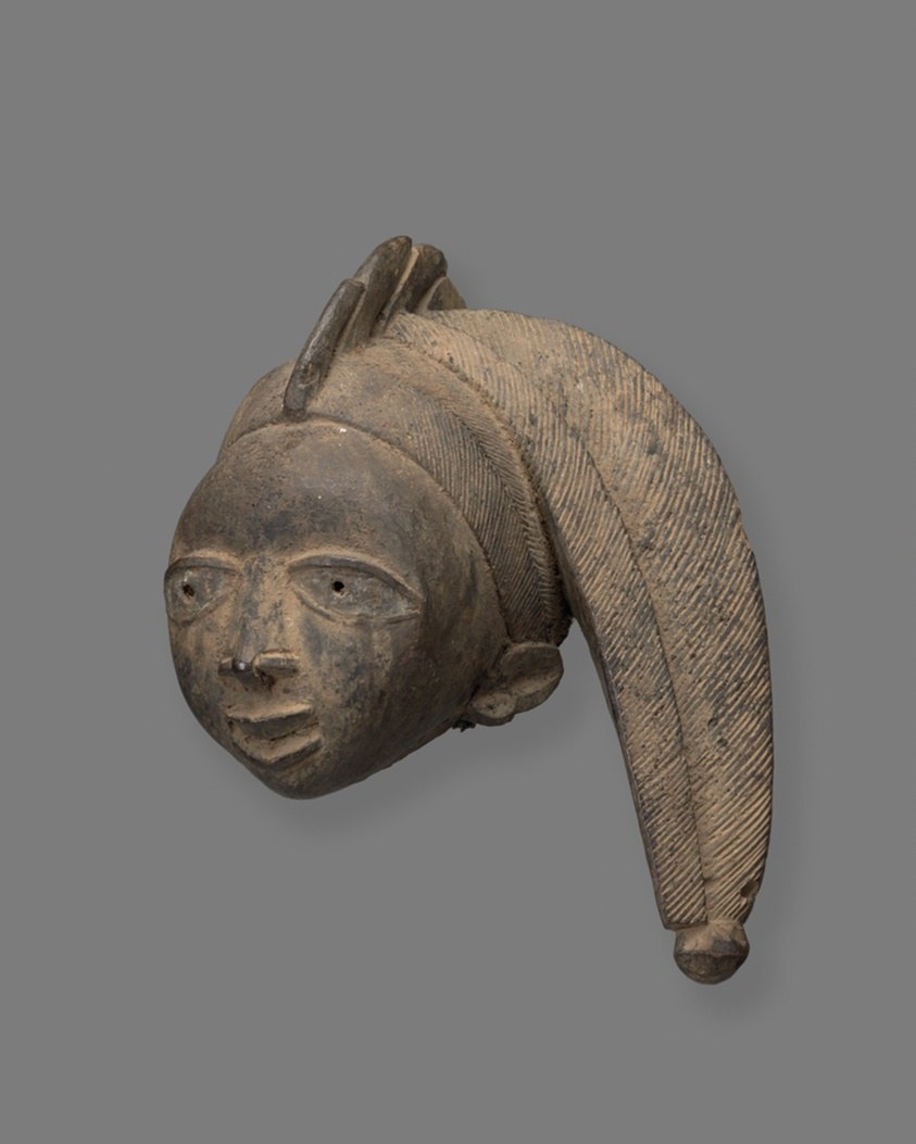 Wooden carved headdress with a stylized human head and curved arm extending downward, with linear surface patterns.