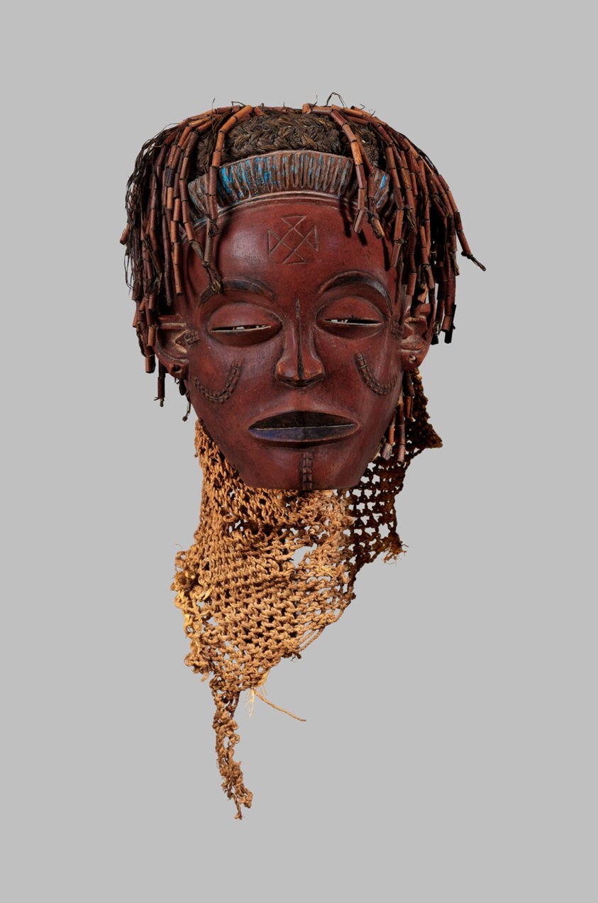 Carved wooden face mask with closed eyes, scarified cheeks, elaborate braided fiber headdress, and fringed collar studded with beads.