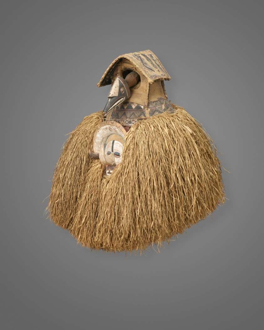 Wooden bird-head helmet mask with incised patterns, topped with a roof-like crest, below which hangs a voluminous skirt of tan raffia fiber. A pale wooden face