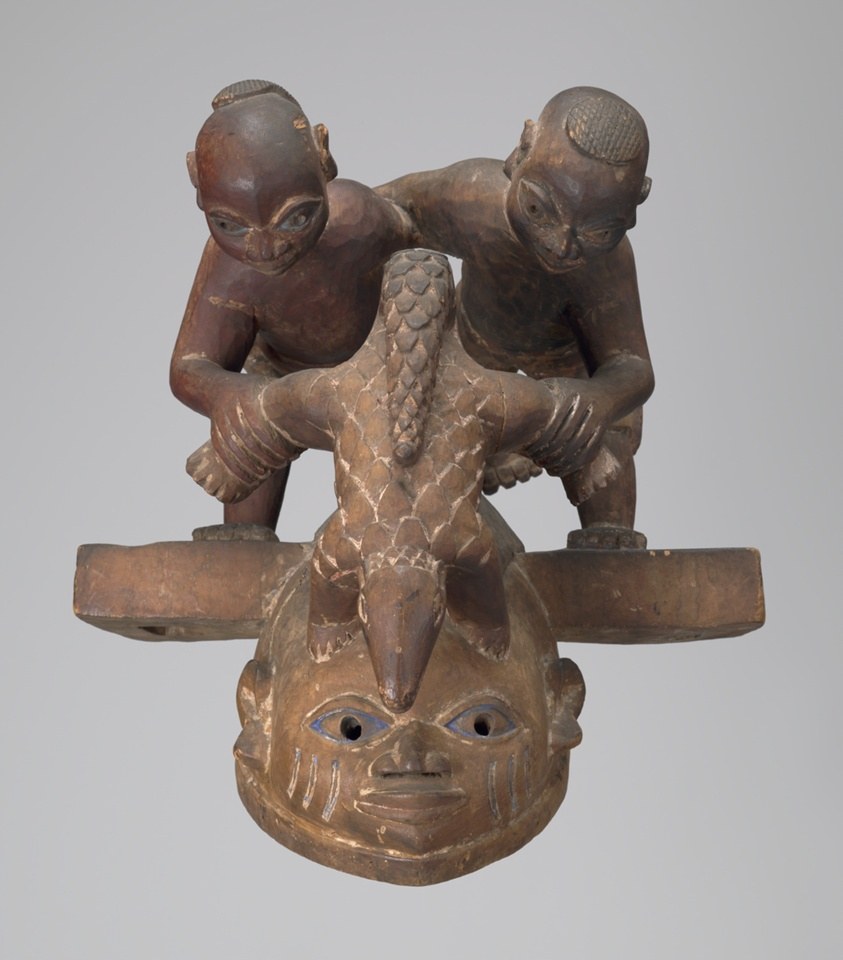 Wooden Yoruba headdress with stacked carved figures: ancestral face below, two small human figures on a horizontal beam, bird with spread wings above.