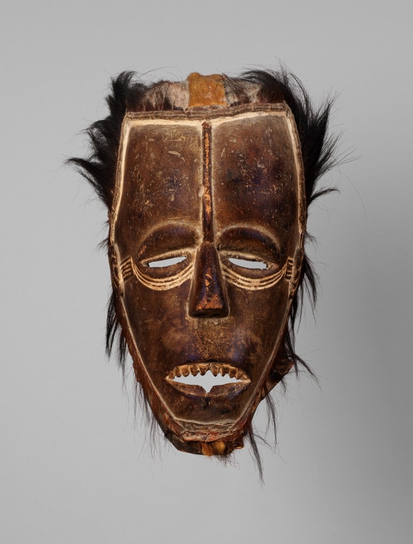 Wooden face mask with kaolin eye and mouth markings, surrounded by long strands of dark monkey fur, from Côte d'Ivoire.