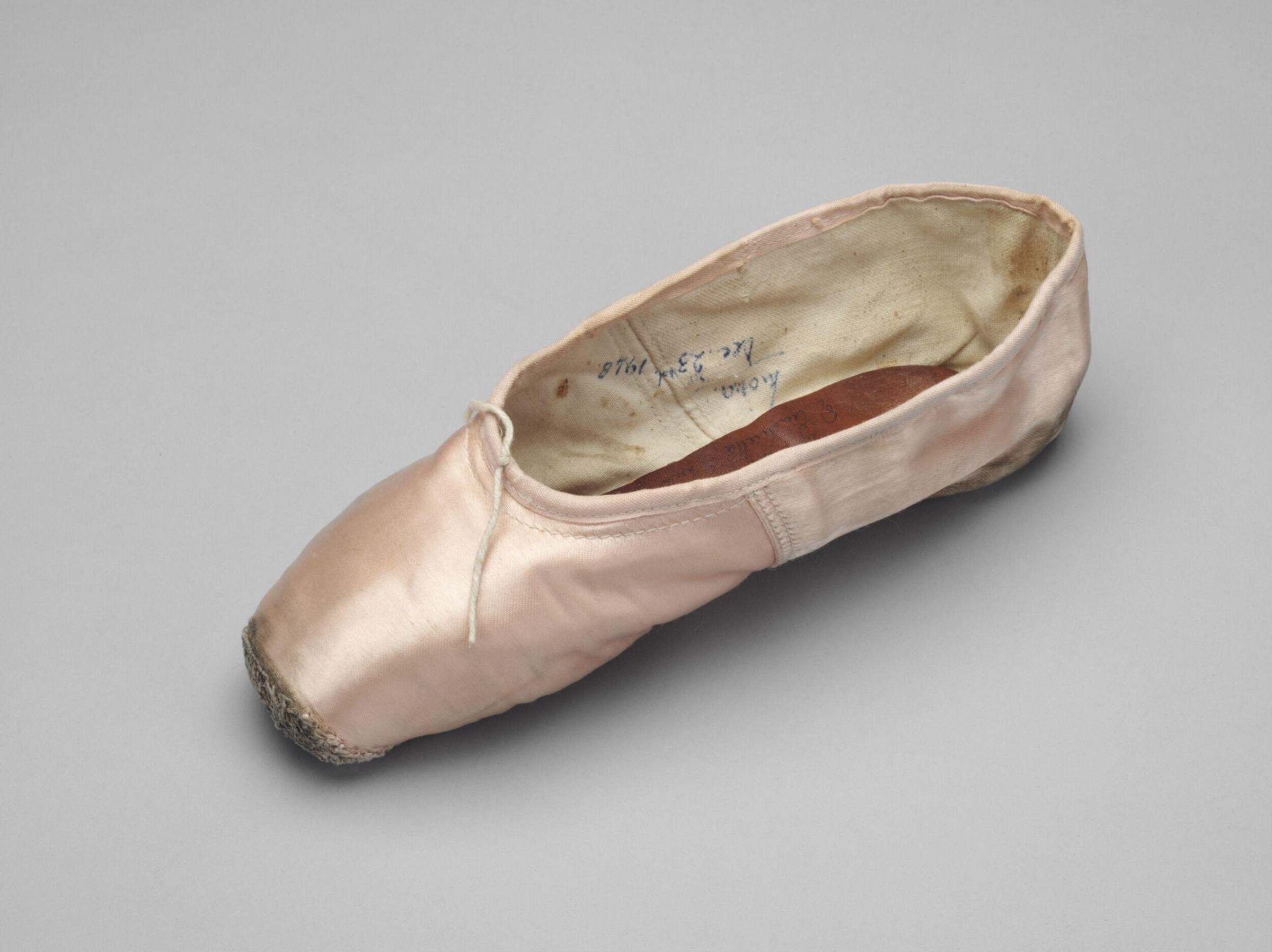 Single pale pink satin pointe shoe shown against grey background, revealing wear on toe box and heel; handwritten inscription visible inside.