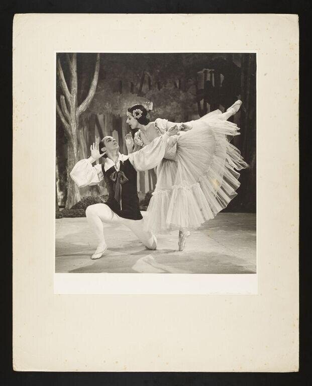 Male dancer in dark jacket kneels beside female dancer in white tutu en pointe, supporting her in a high extension with dramatic lean backward.