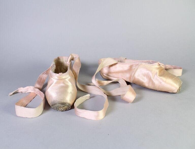 Two worn pink pointe shoes on gray background, ribbons extended, showing creased satin and marked soles.