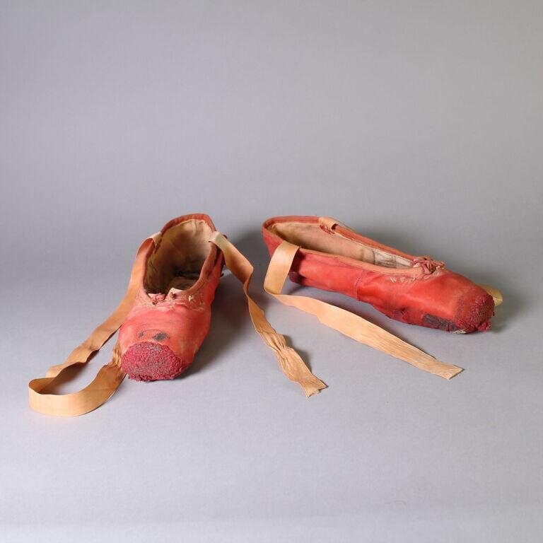Two worn coral-pink satin pointe shoes with pale ribbons, shown against a grey background to display their deterioration and use.