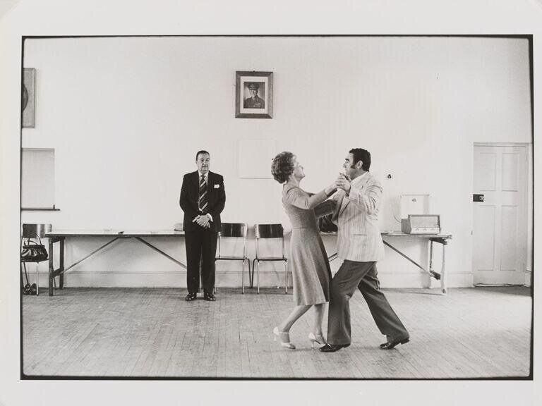 Ballroom dance instructor Ted van Rensburg watches a couple execute a closed frame waltz or foxtrot in an institutional hall, Boksburg, 1980.