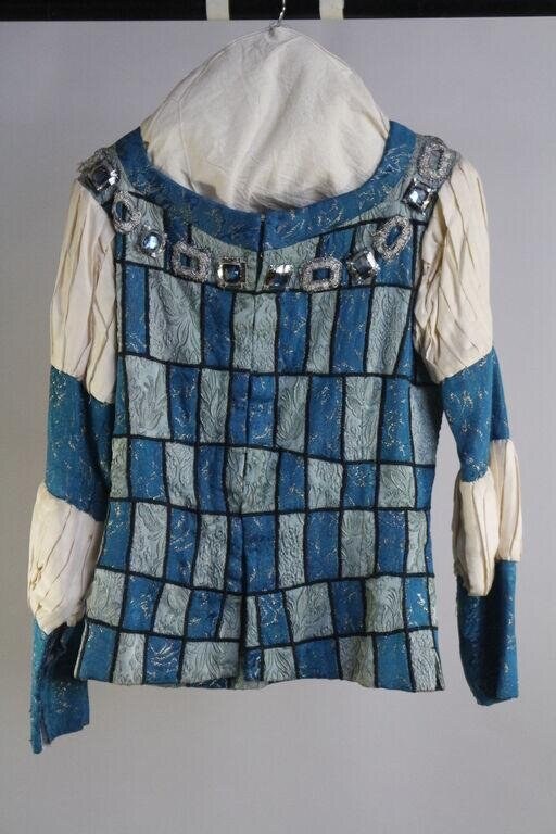 Back view of a sleeveless tunic costume with alternating blue and pale green patchwork squares, dark bound edges, and cream-colored sleeves with decorative