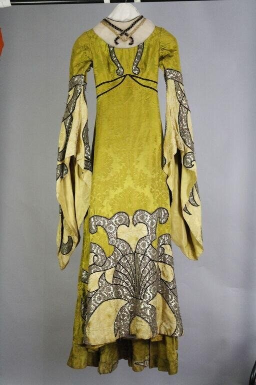 Yellow-green linen tunic with ornamental trim, displayed on a dress form. Decorative chains drape the shoulders; elaborate woven or embroidered bands frame the