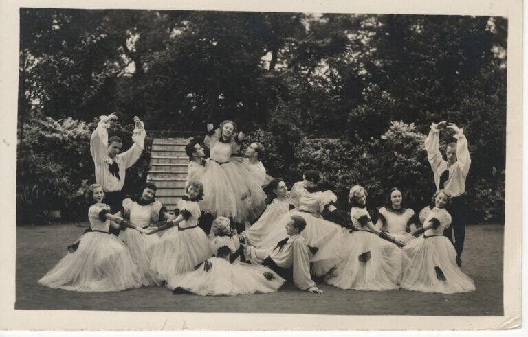 Fifteen young women in white drapery arranged in a tableau on a garden lawn, posed with raised arms and flowing skirts, photographed in soft focus black and