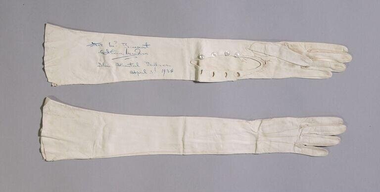 Two long cream-colored fabric gloves or sleeves, arranged flat. Upper piece bears blue handwritten text dated 1934.