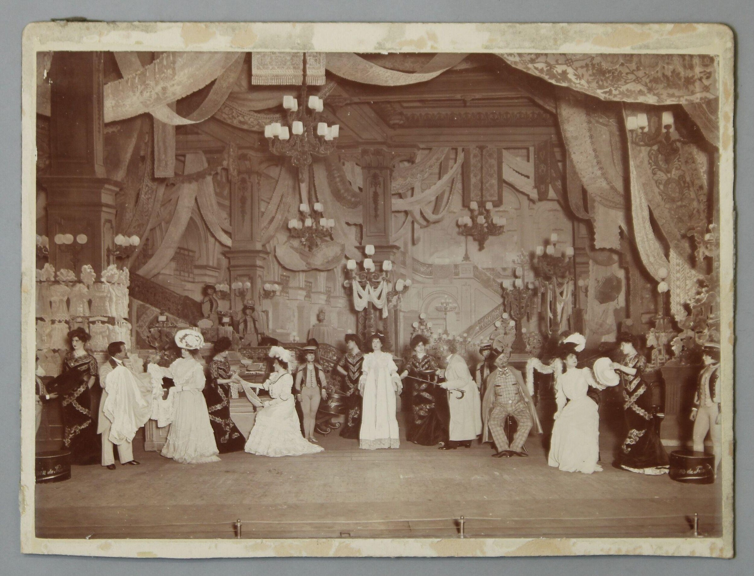 Sepia photograph of a formal ball scene with twelve figures in evening dress arranged on a stage beneath ornate classical architecture and hanging chandeliers.