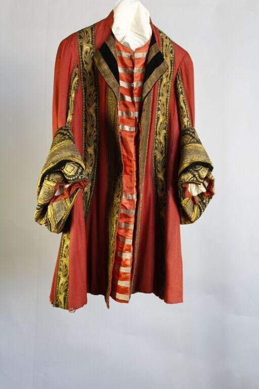 Red silk theatrical costume coat with gold and black embroidered cuffs depicting winged creatures, laid on a dress form.