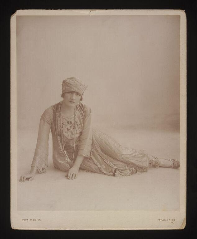 Female dancer in ornate beaded gown kneeling on a stage floor, head tilted back in a dreamy pose, one arm extended forward.