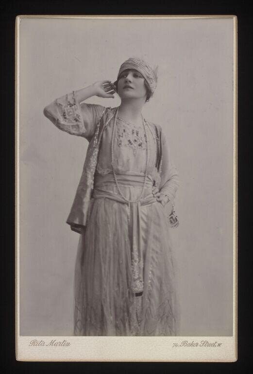 Woman in Edwardian costume and embroidered cap stands gazing upward, left arm raised to her head, draped in a flowing gown.